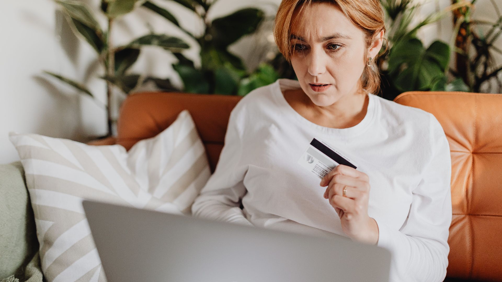 A woman sitting on a couch shopping online with a credit card and laptop in a cozy living room.