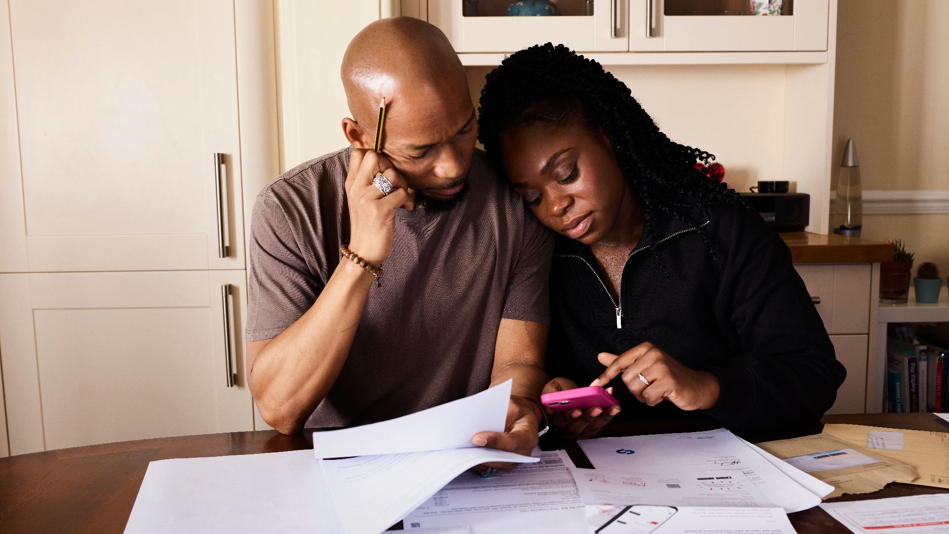 A couple sits at a table managing domestic finances, evaluating documents and using a smartphone.