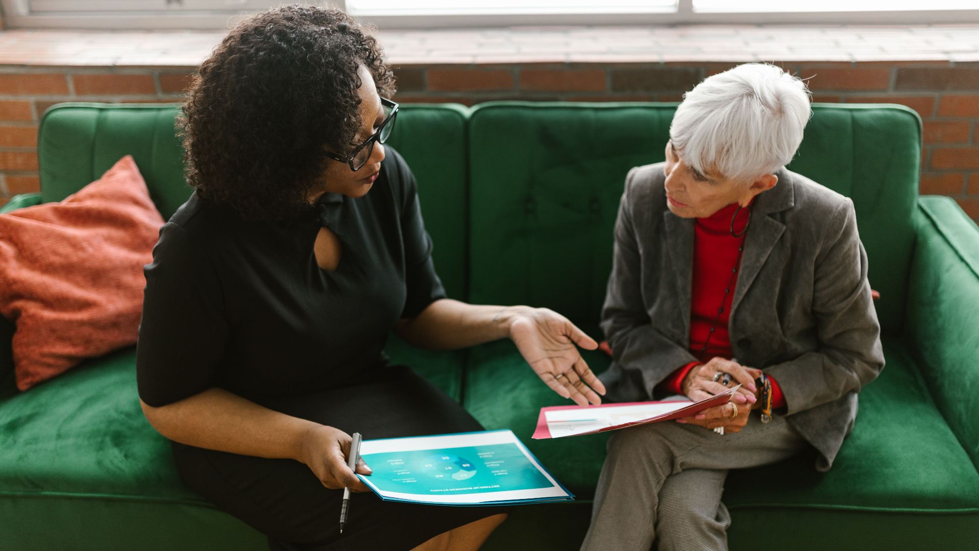 Two women in a business meeting discuss documents while sitting on a green sofa.