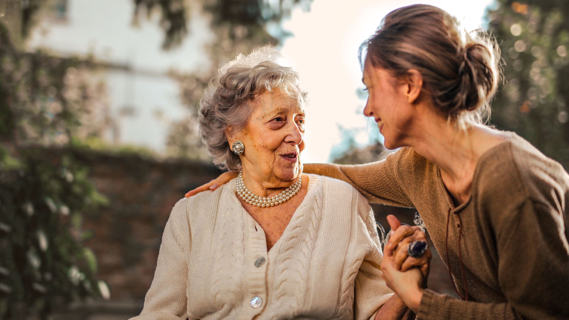 Low angle of cheerful aged lady and adult woman in casual clothes holding hands and smiling at each other while having meeting on yard against blurred green trees and facade of country house in suburb