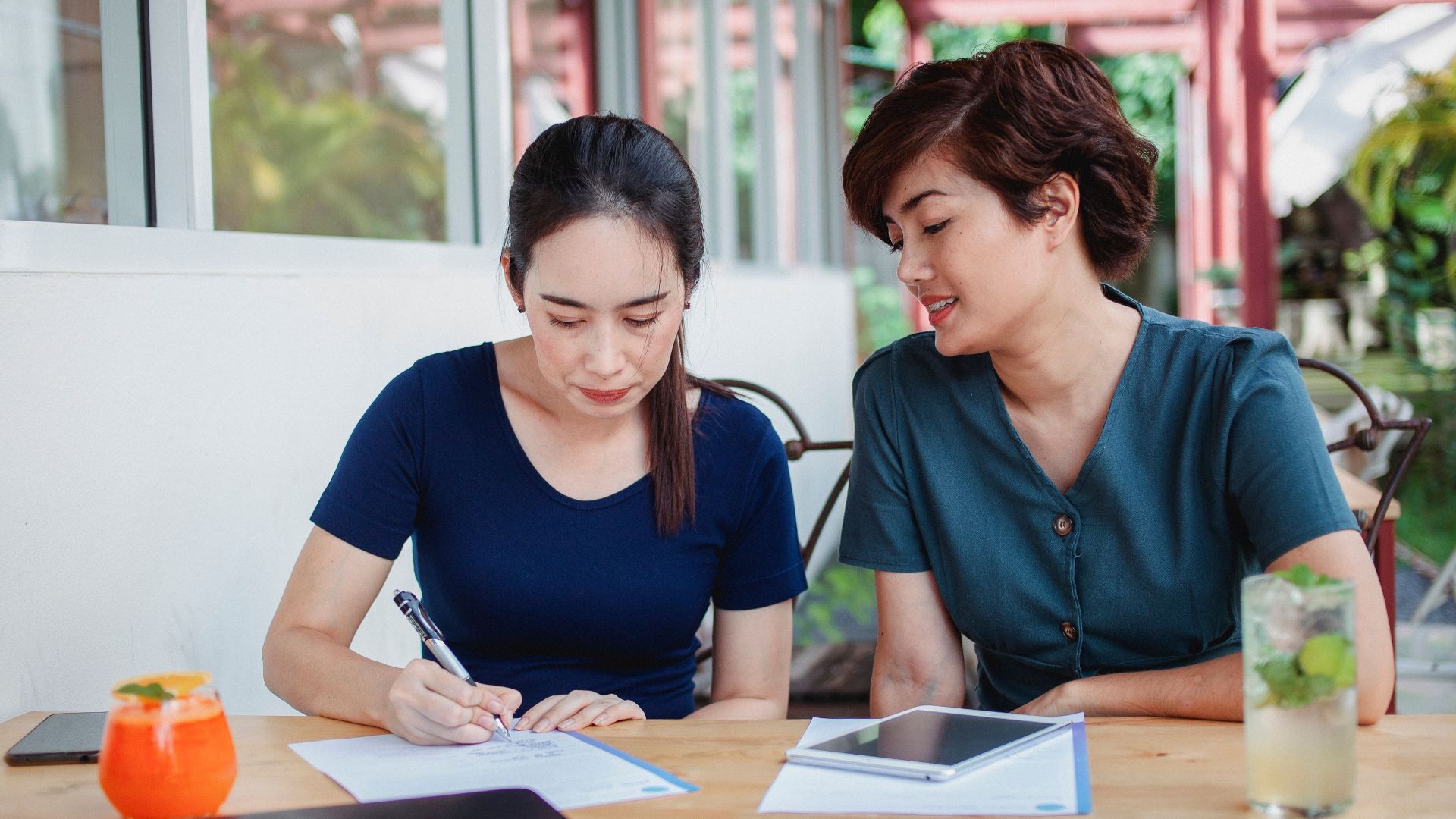 Young ethnic female partners sitting at table with gadgets and documents and signing contract on terrace of cafe
