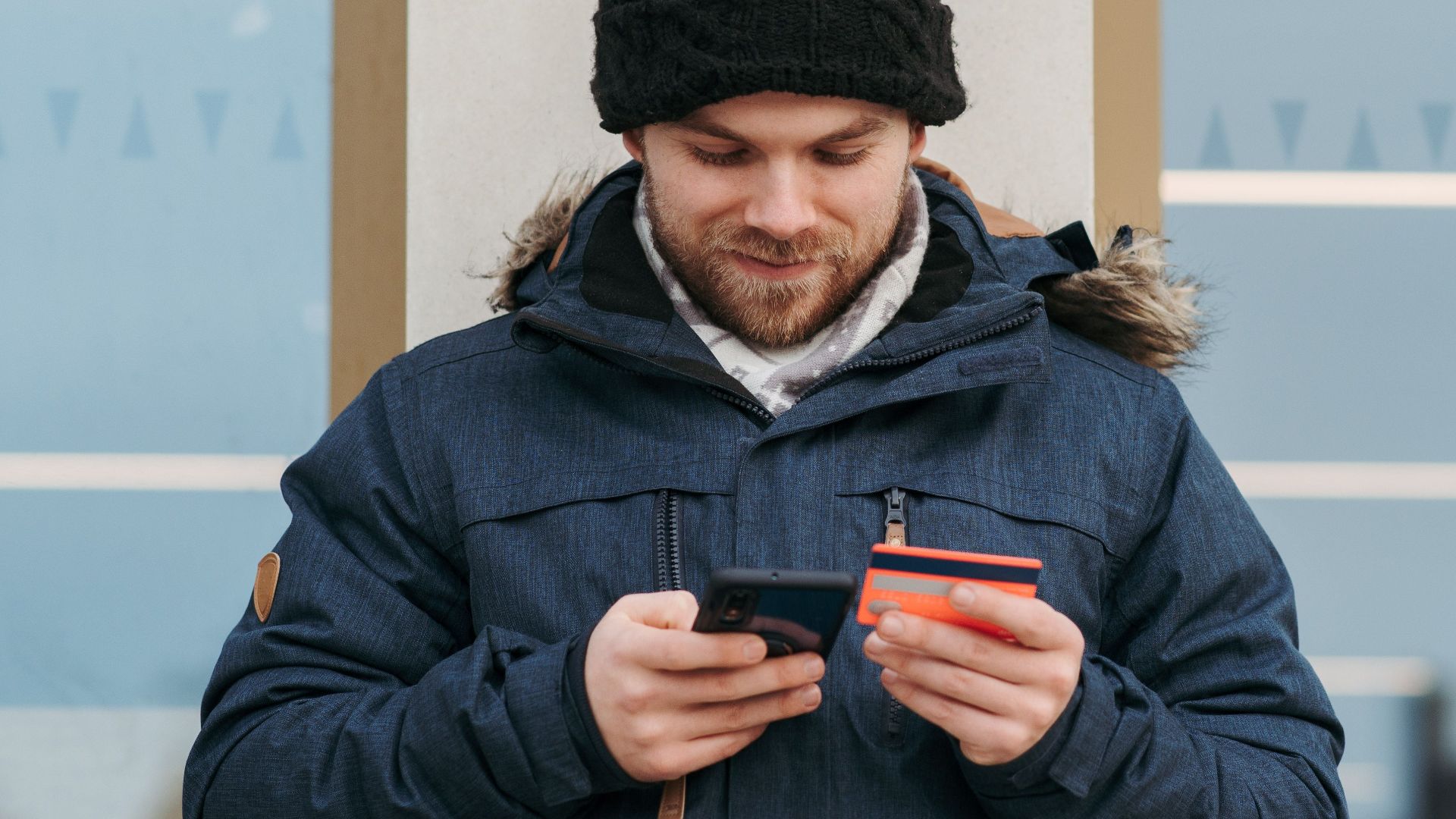 Positive man in warm clothes standing near building and entering credentials of credit card on mobile phone in daylight