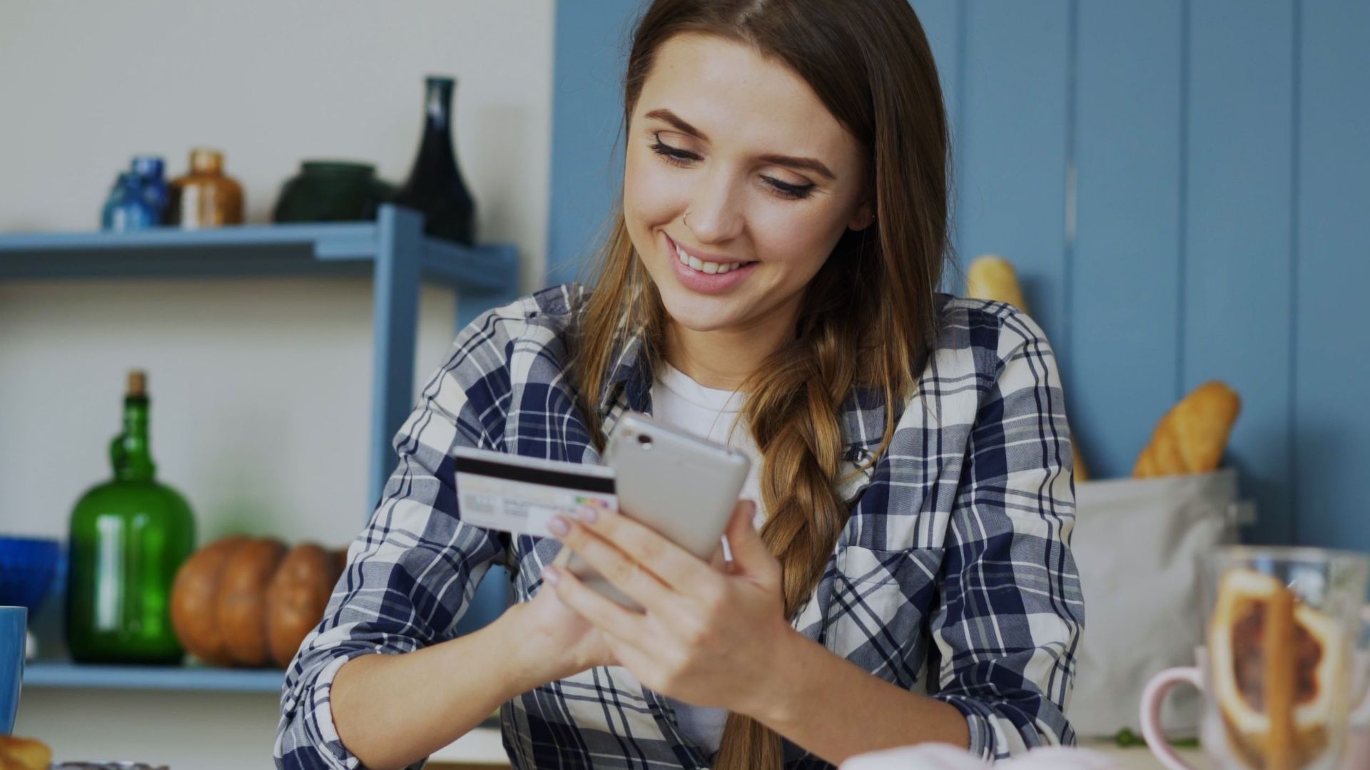 Smiling woman using smartphone and credit card for online shopping in a cozy indoor kitchen.