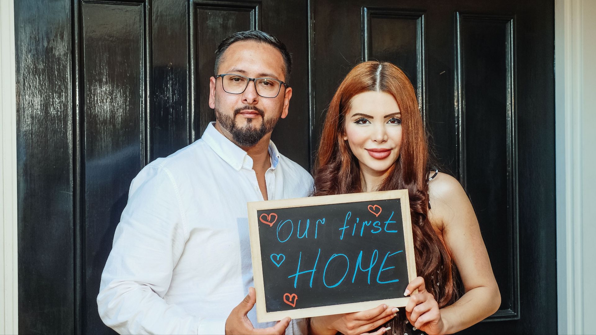 A joyful couple stands at their new home's door, holding a chalkboard saying 'Our First Home'.