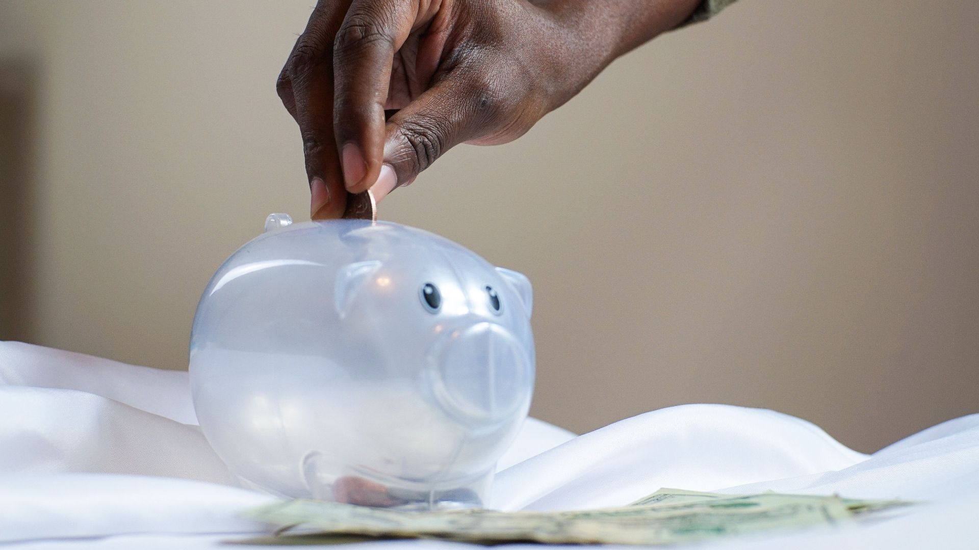 Close-up of a person's hand placing coins into a transparent piggy bank to save money.