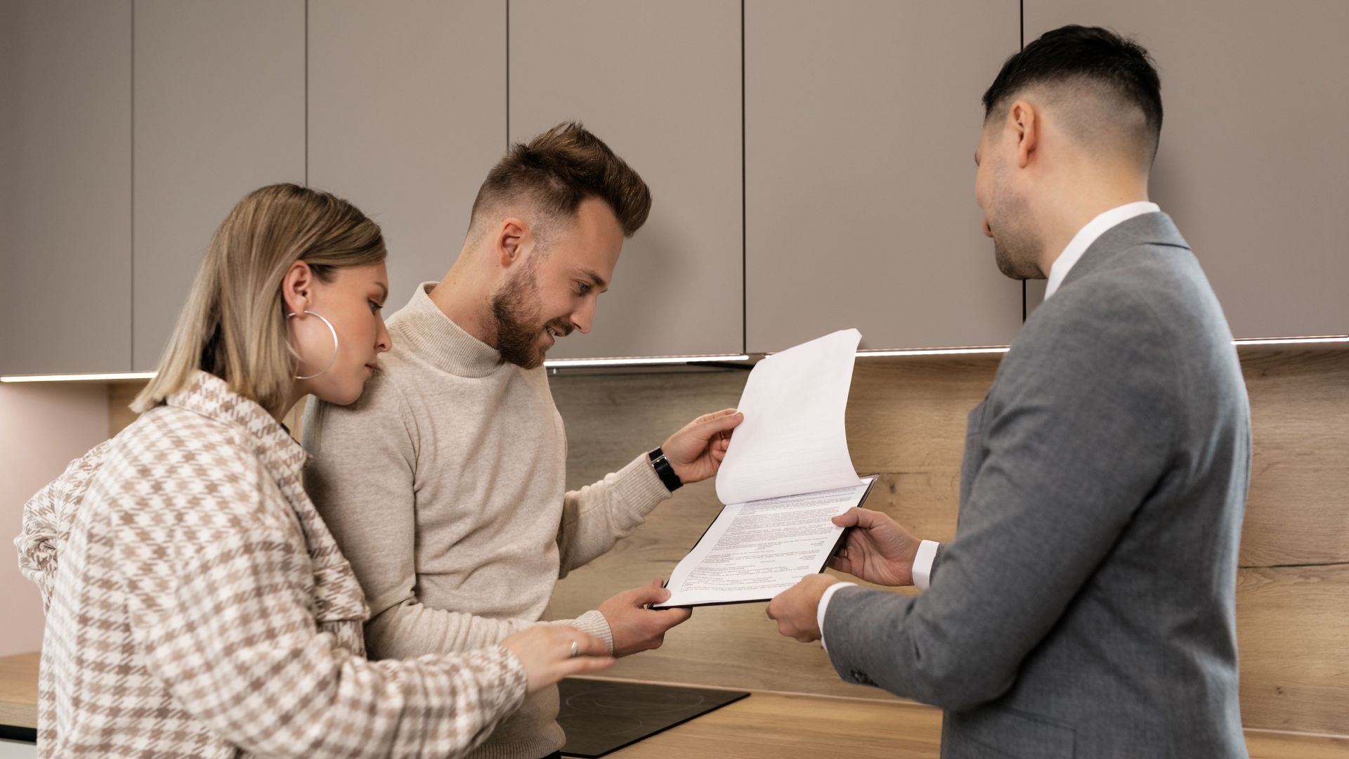 A couple reviews real estate documents with an agent in a modern indoor setting, discussing a potential property purchase.