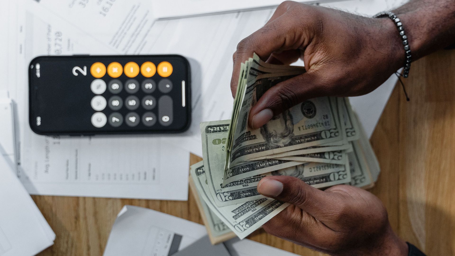 Person counting dollar bills over documents with a smartphone calculator on the desk.