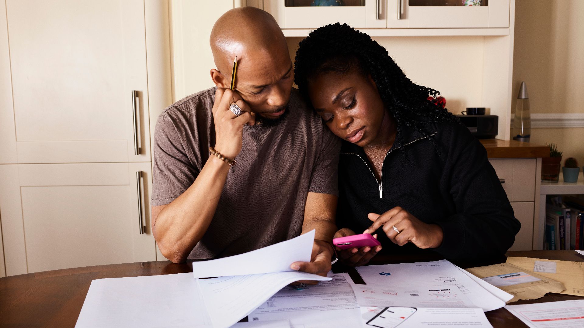 A couple sits at a table managing domestic finances, evaluating documents and using a smartphone.