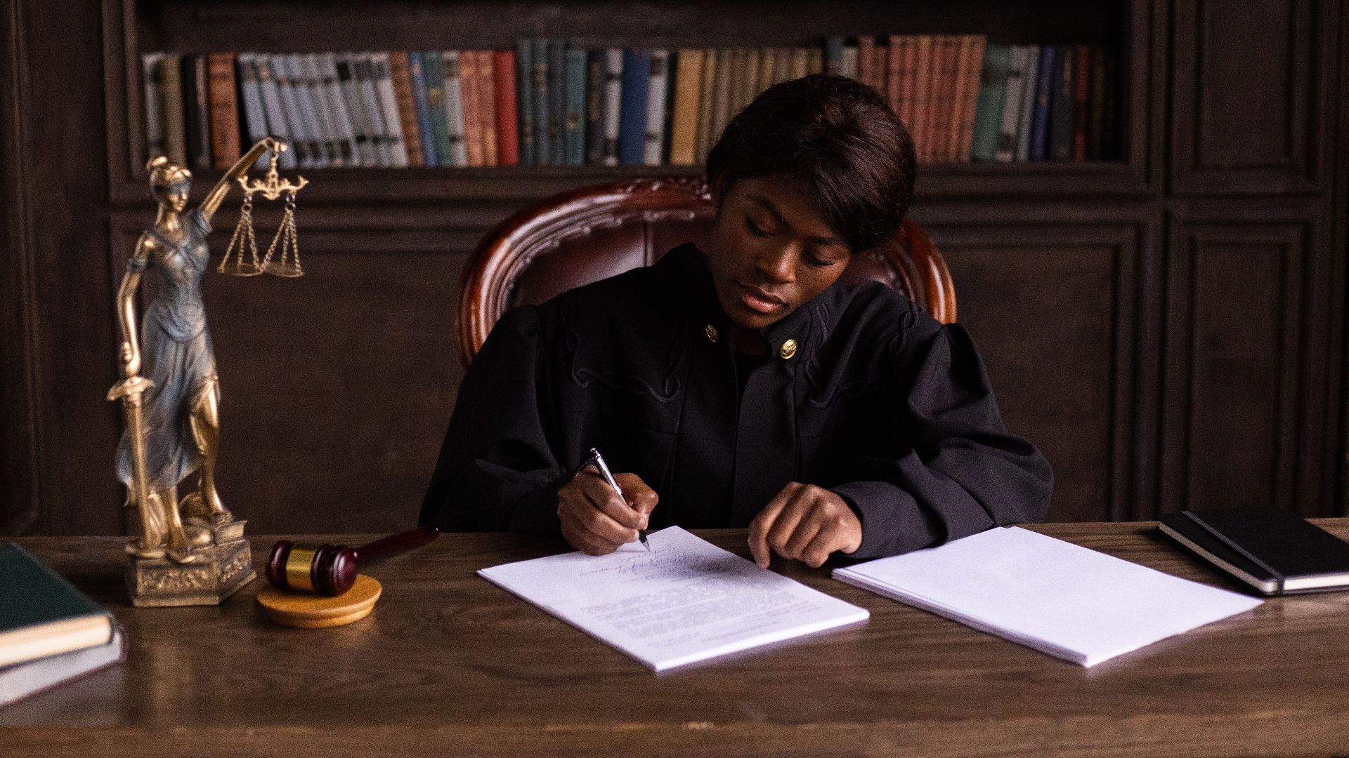 A judge in robes writing on a document at a desk in an office library with law books.