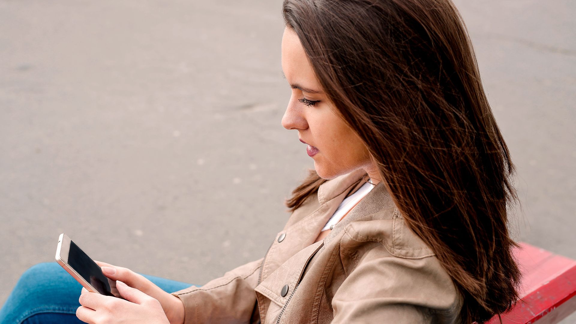A woman in a brown jacket using a smartphone while sitting on a bench outdoors.