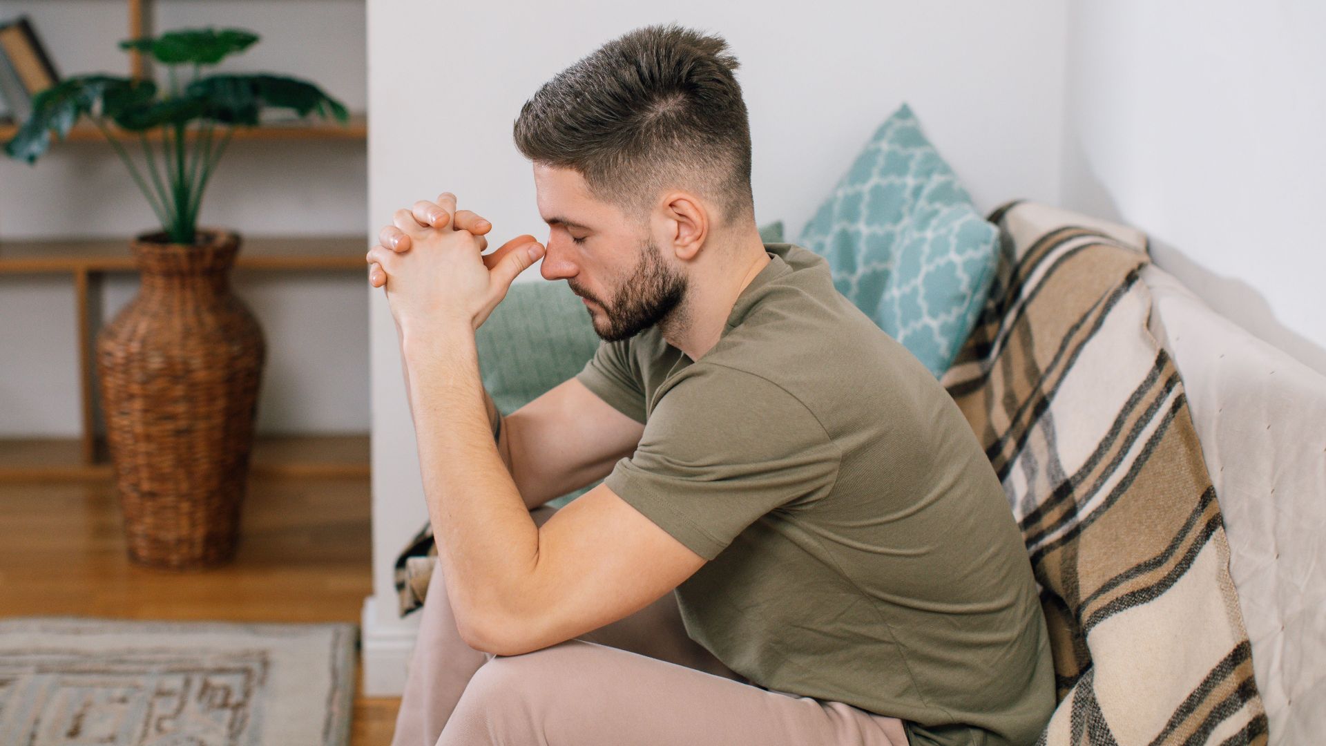 A young man with clasped hands, sitting thoughtfully on a sofa in a cozy living room.