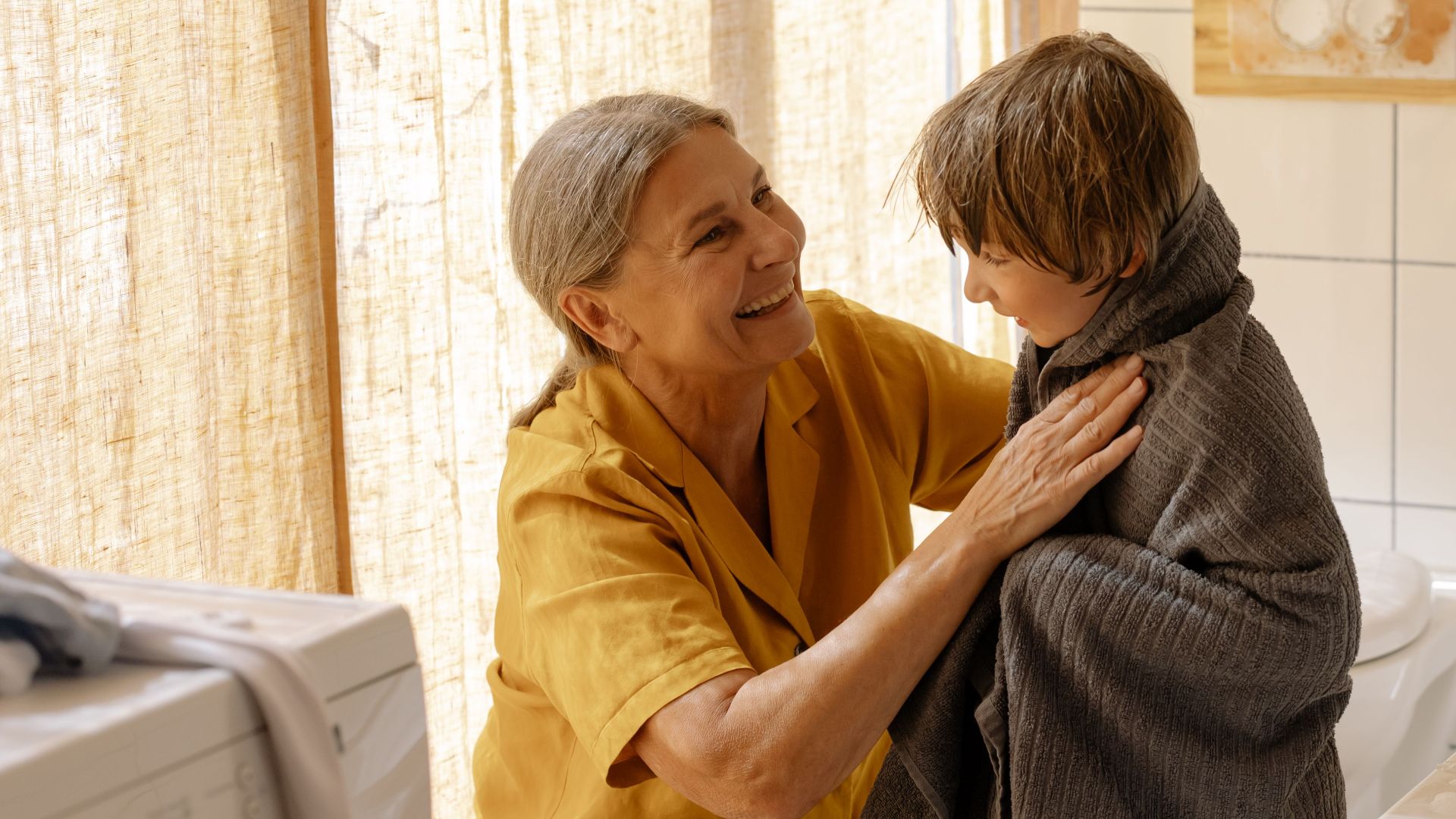 Grandmother drying her grandson with a towel in a warm, sunlit bathroom.