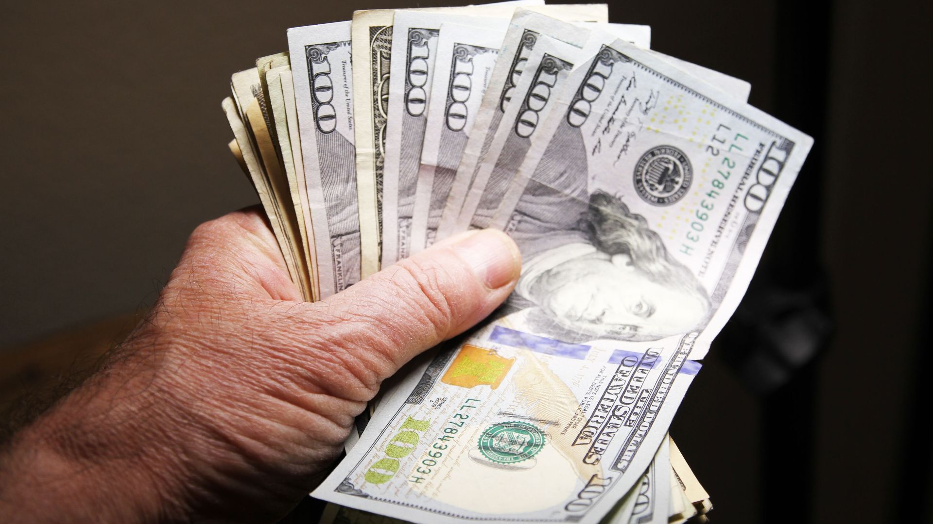 Close-up of a hand holding multiple hundred dollar bills on a dark background.