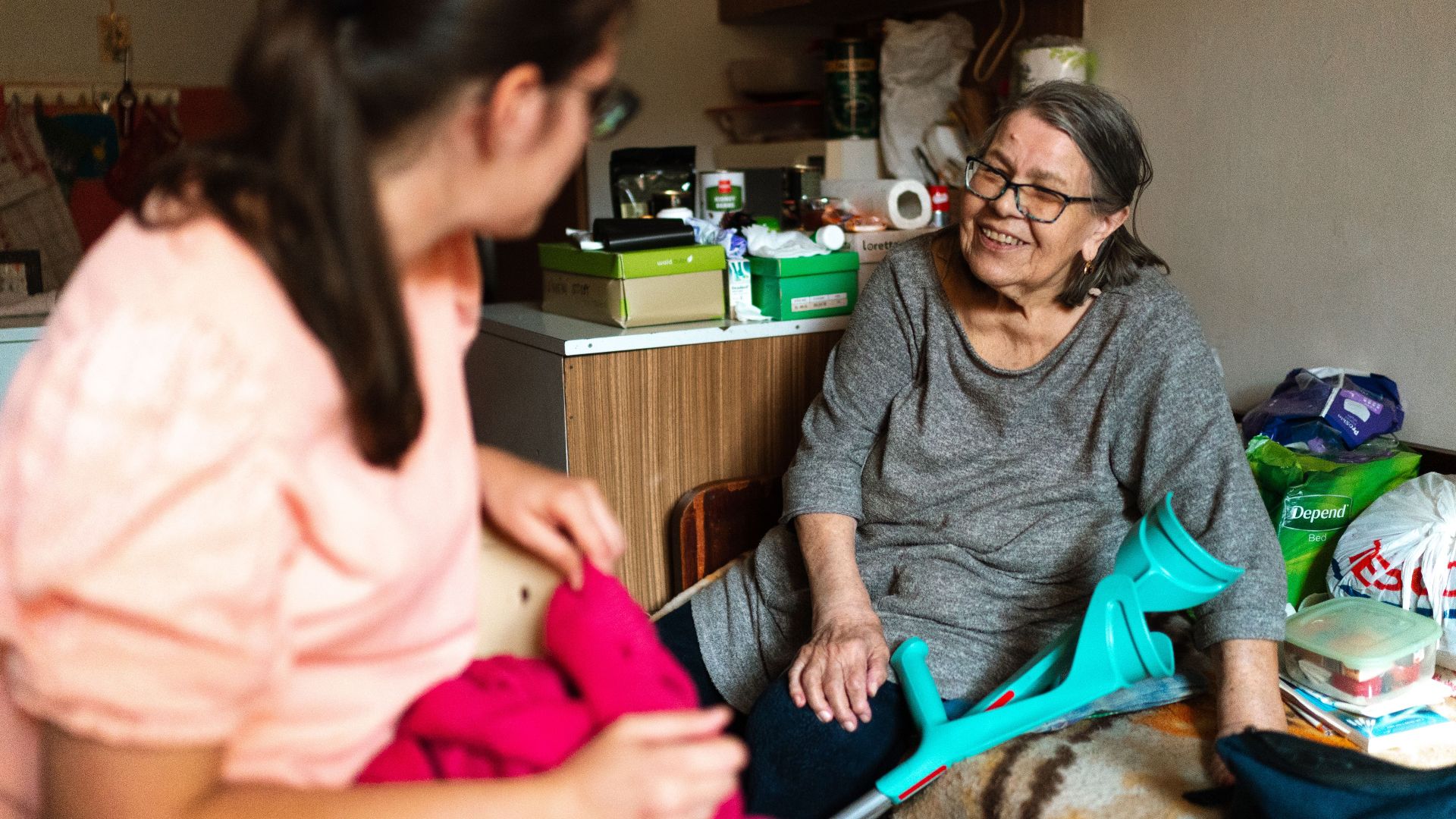A caregiver helps a senior woman with a crutch in a cozy Prague home setting.