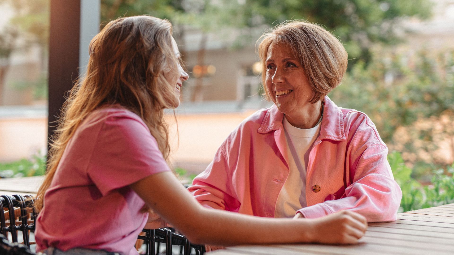 Two women smiling and chatting at a wooden table outdoors, depicting friendship and connection.
