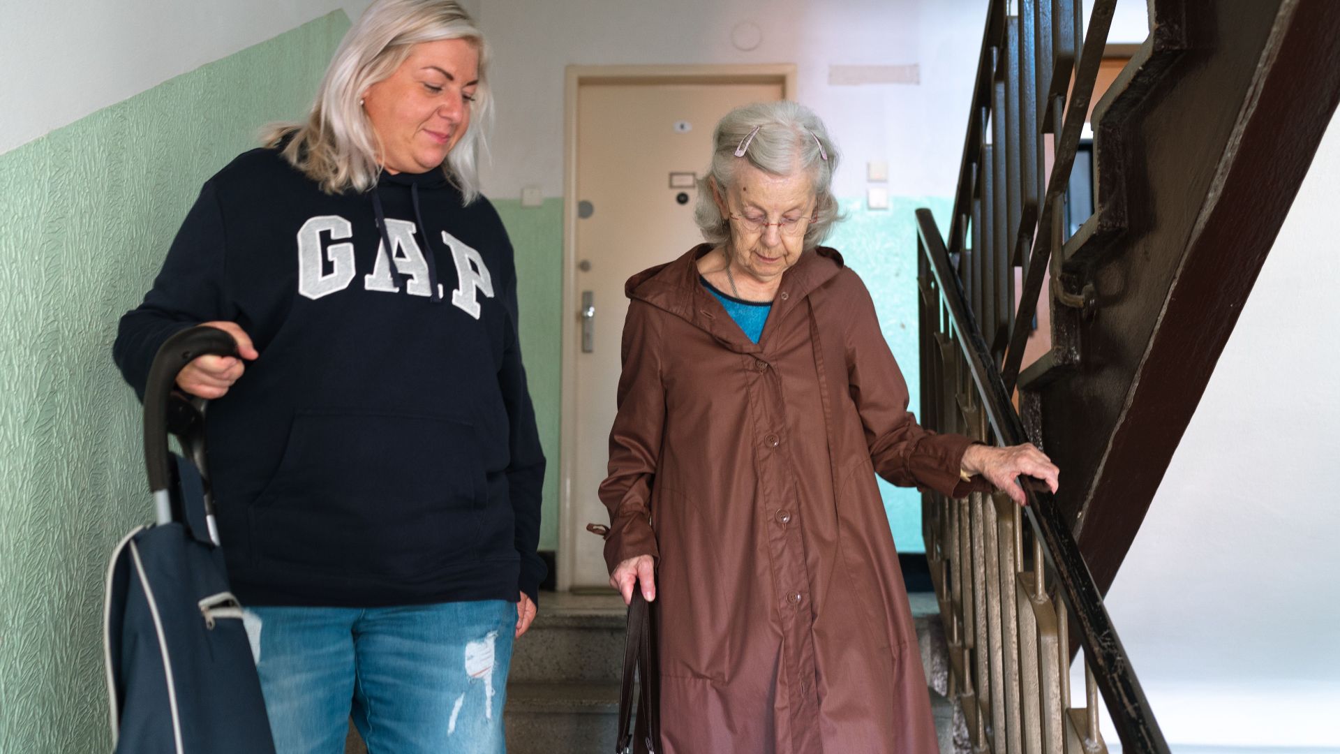 A senior woman assisted by a caregiver walking down indoor stairs in Prague.