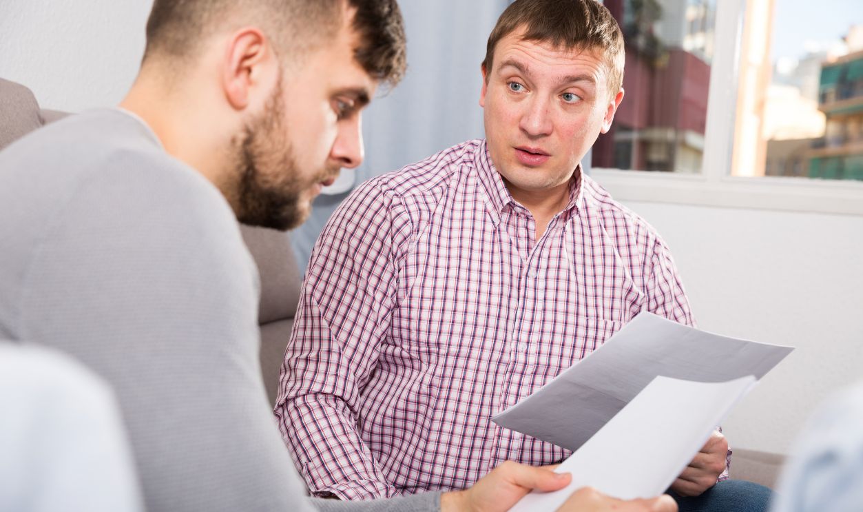 Upset man sitting on sofa at home with friend, looking worriedly at papers