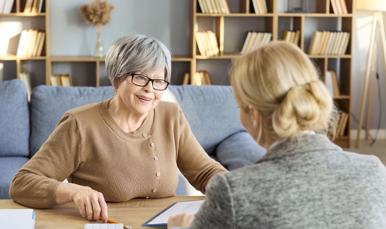 Senior woman and advisor in consultation. At home client reviews paperwork with an advisor, discussing savings insurance and estate planning with a calculator. Clear concept of financial help.