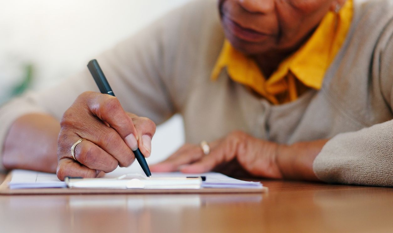 Senior woman, hands and writing agreement on contract, form or application for retirement plan or insurance at home. Closeup of elderly female person signing documents or paperwork on table at house
