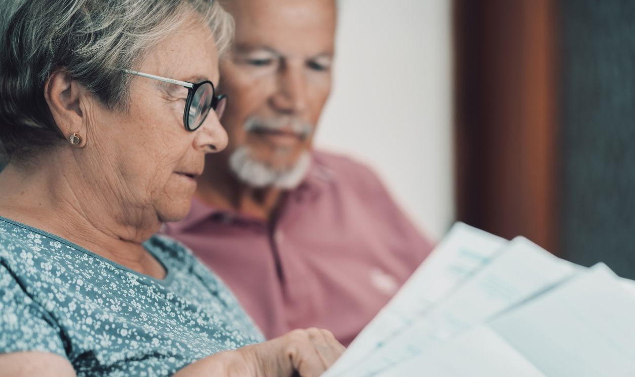 Senior couple sits in their living room reading and discussing bills and paperwork, using a laptop for online banking and retirement planning. The scene conveys partnership, care, and household financial management.
