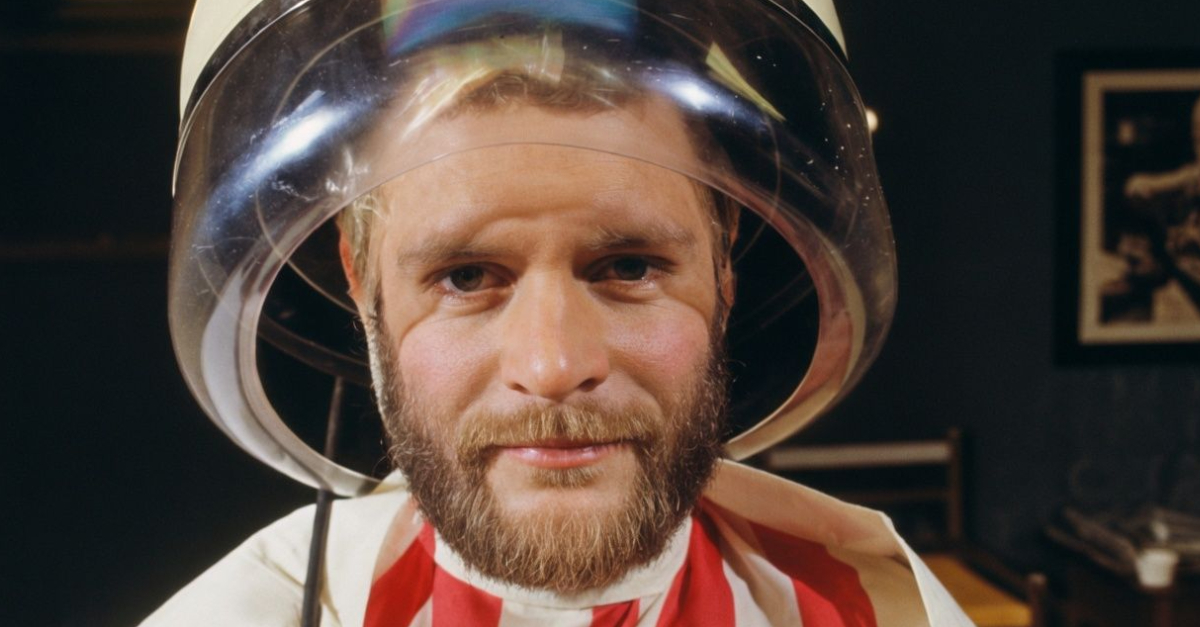 A man is sitting comfortably drying his hair under a drying hood. 1976