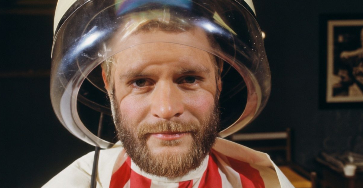 A man is sitting comfortably drying his hair under a drying hood. 1976