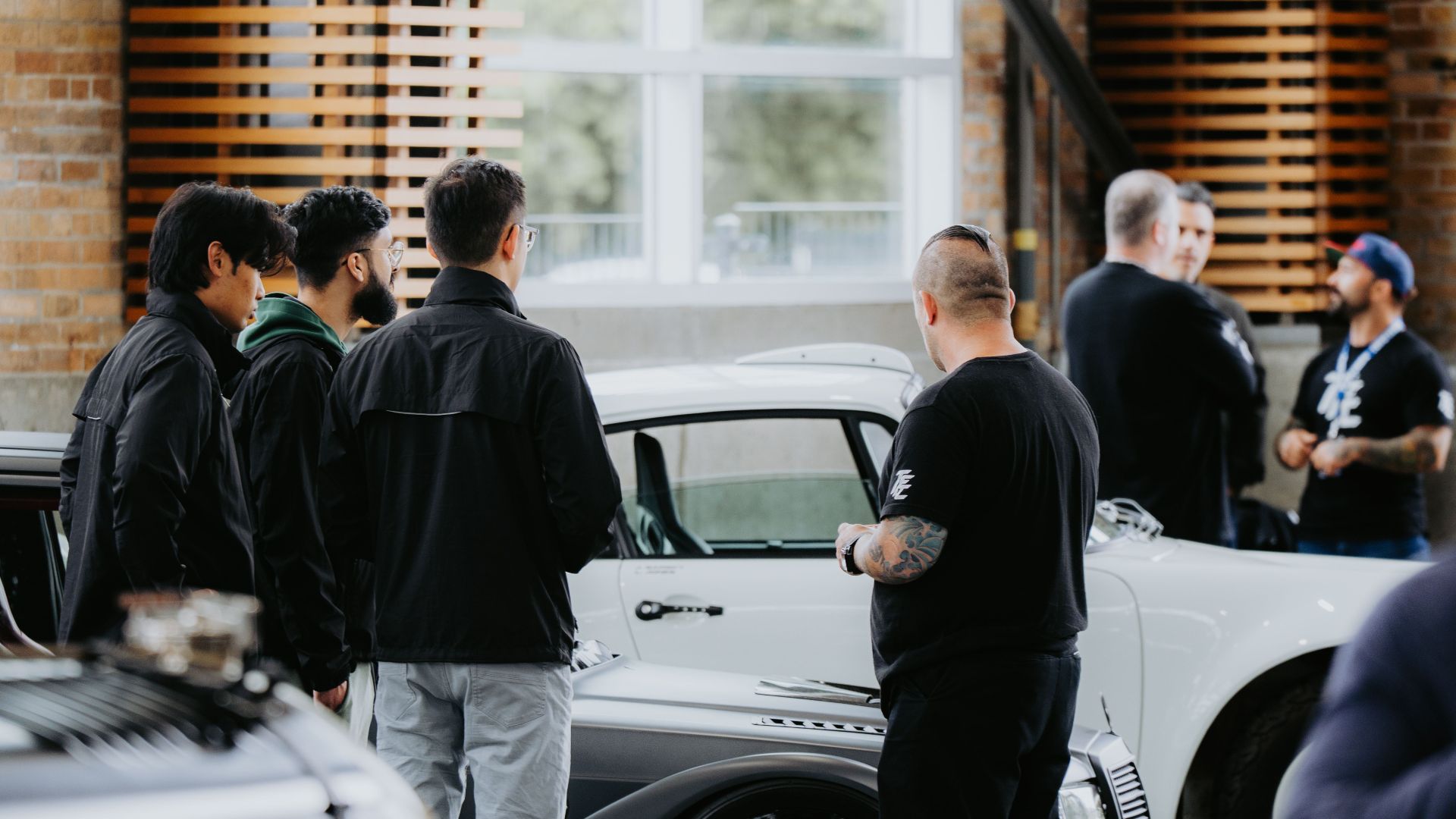 Men gather around vintage cars in an indoor setting, discussing automotive designs.