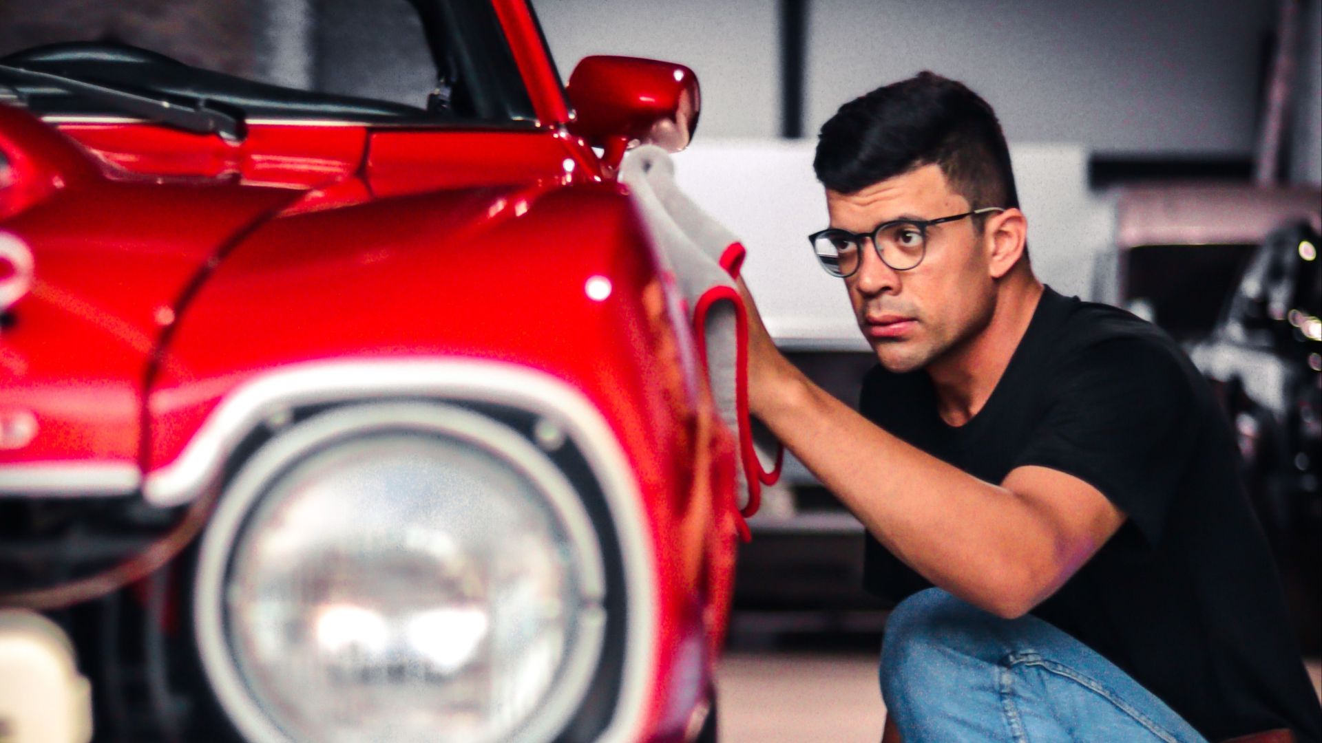 A man kneels to inspect a classic red car in a garage setting, emphasizing detail and care.