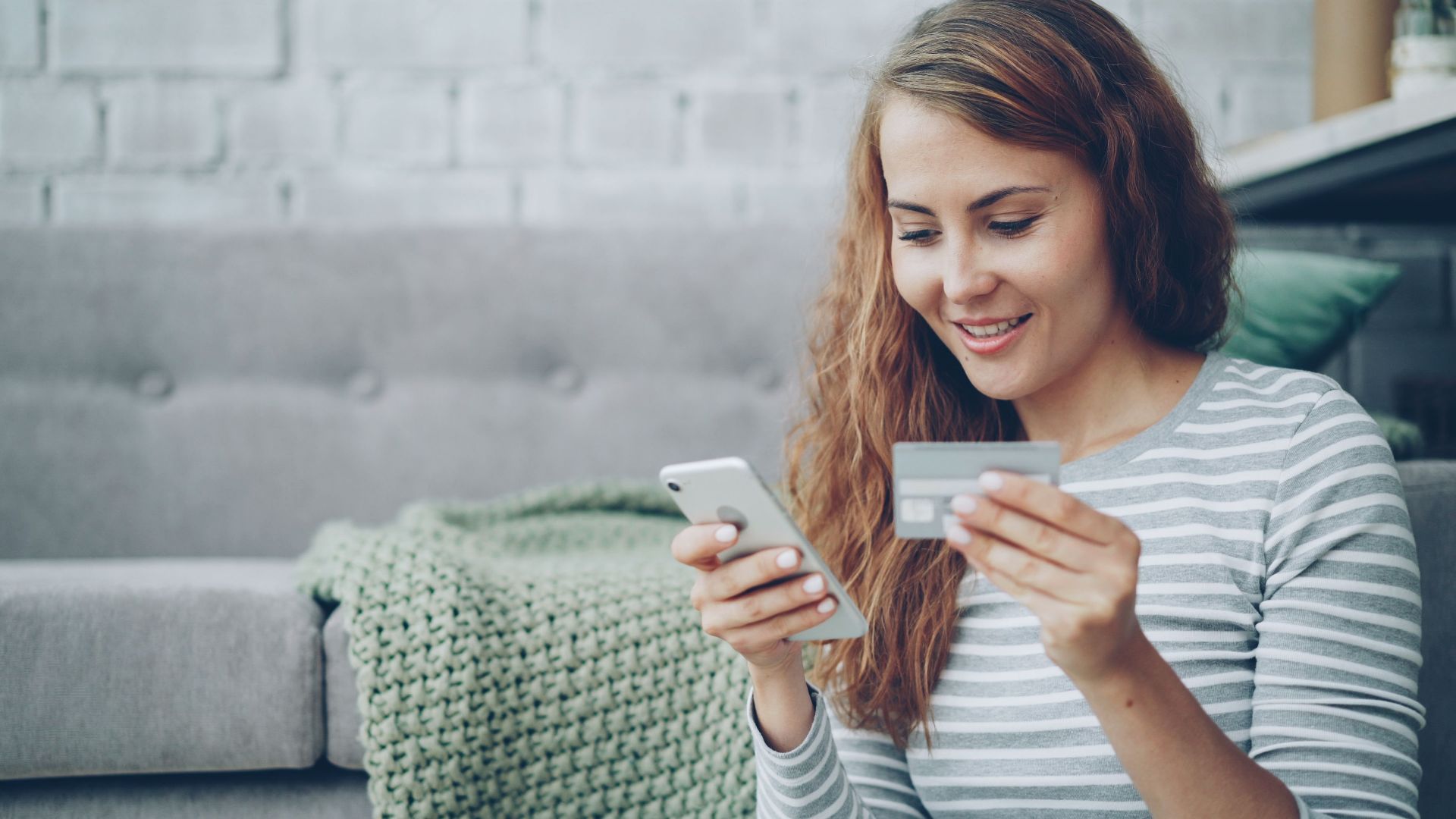 A smiling woman uses her smartphone and credit card for online shopping in a cozy living room.