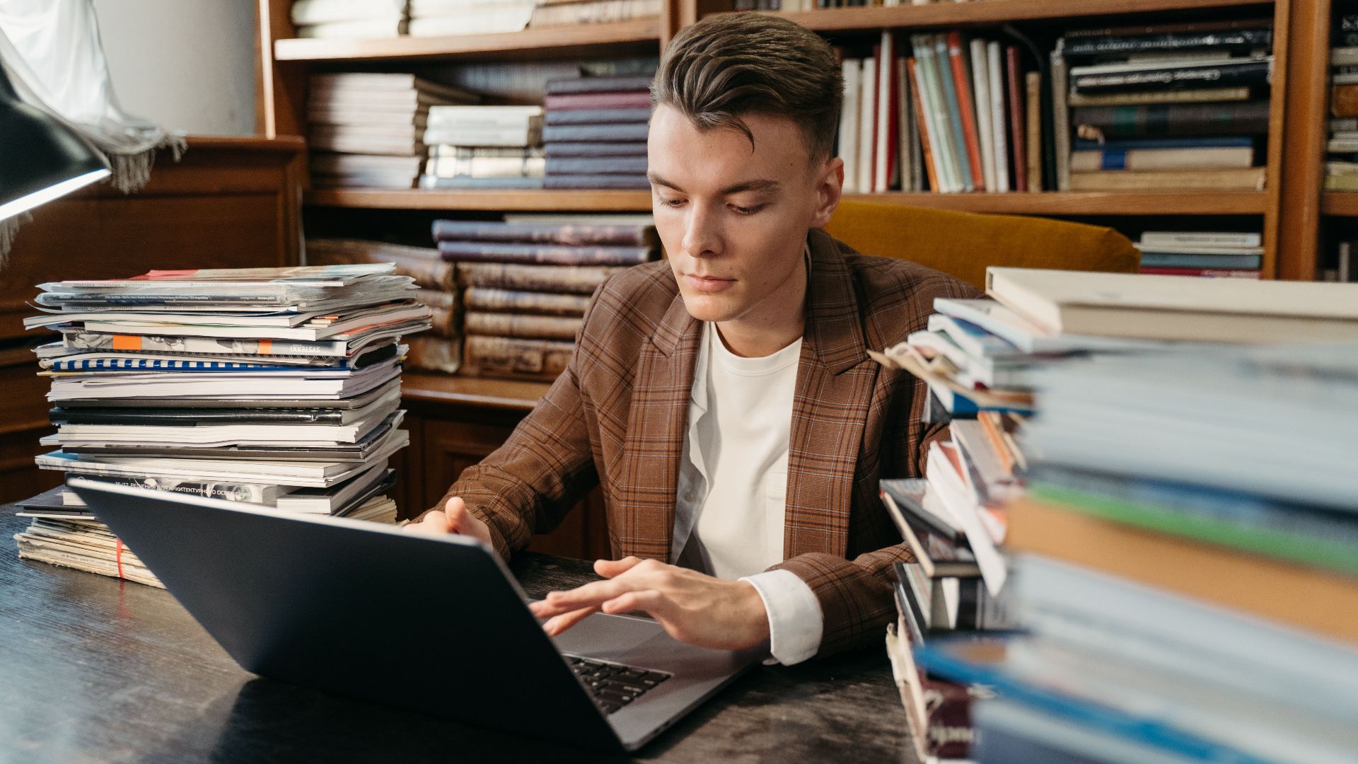 A focused young man studies on a laptop amidst stacks of books in a cozy library setting.