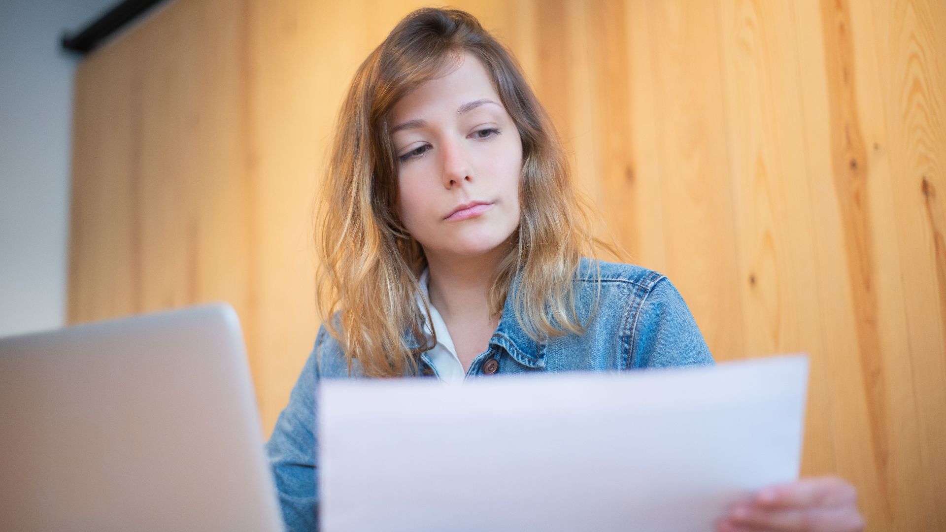 Businesswoman in a denim jacket analyzing documents at a wooden table in a cozy workspace.
