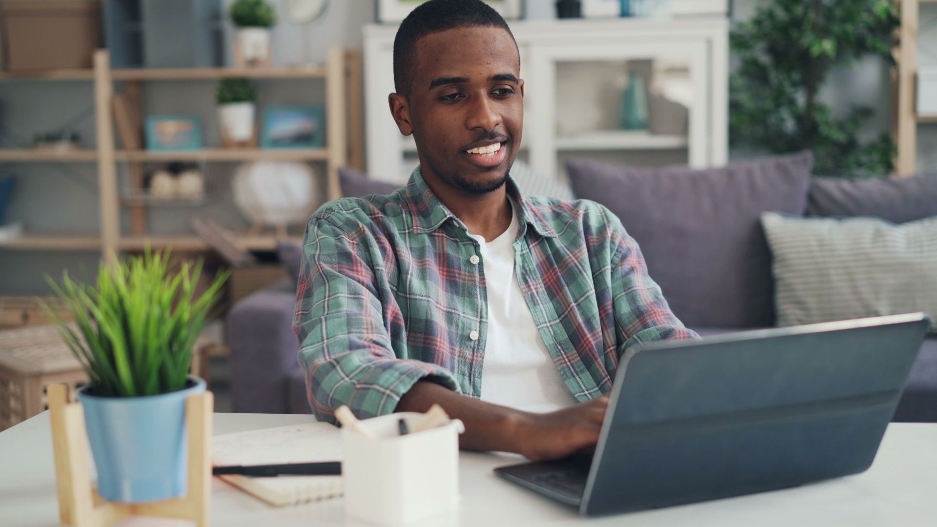 Young man smiling while using laptop in a modern home office setting.