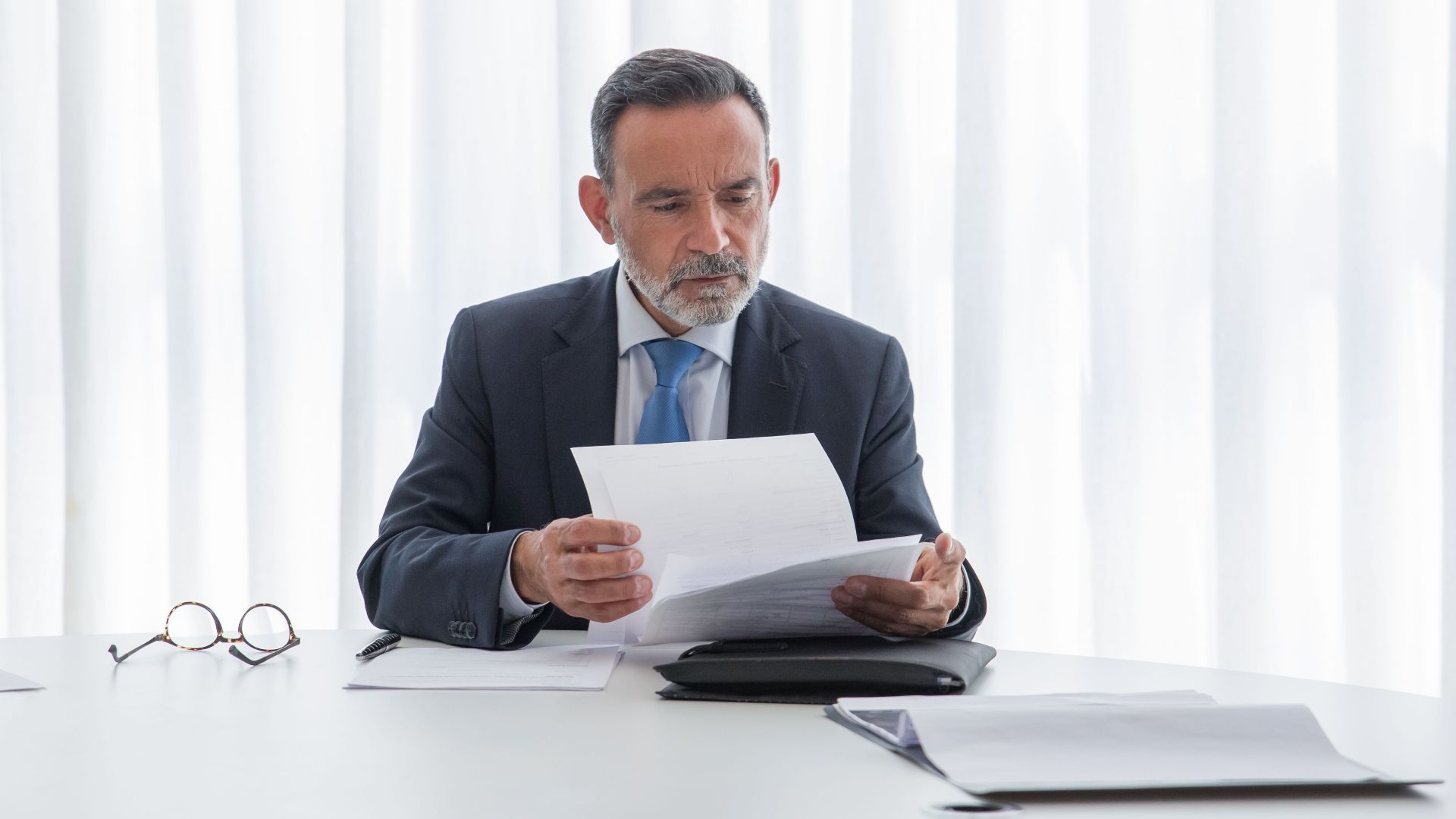 Elderly businessman focusing on reading documents at a table in a bright office setting.