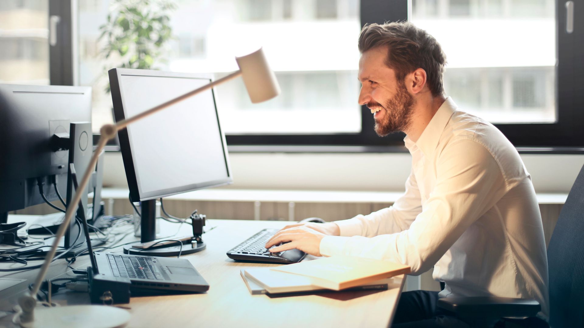 A man smiling while working at an office desk with a computer and natural daylight streaming in through large windows.