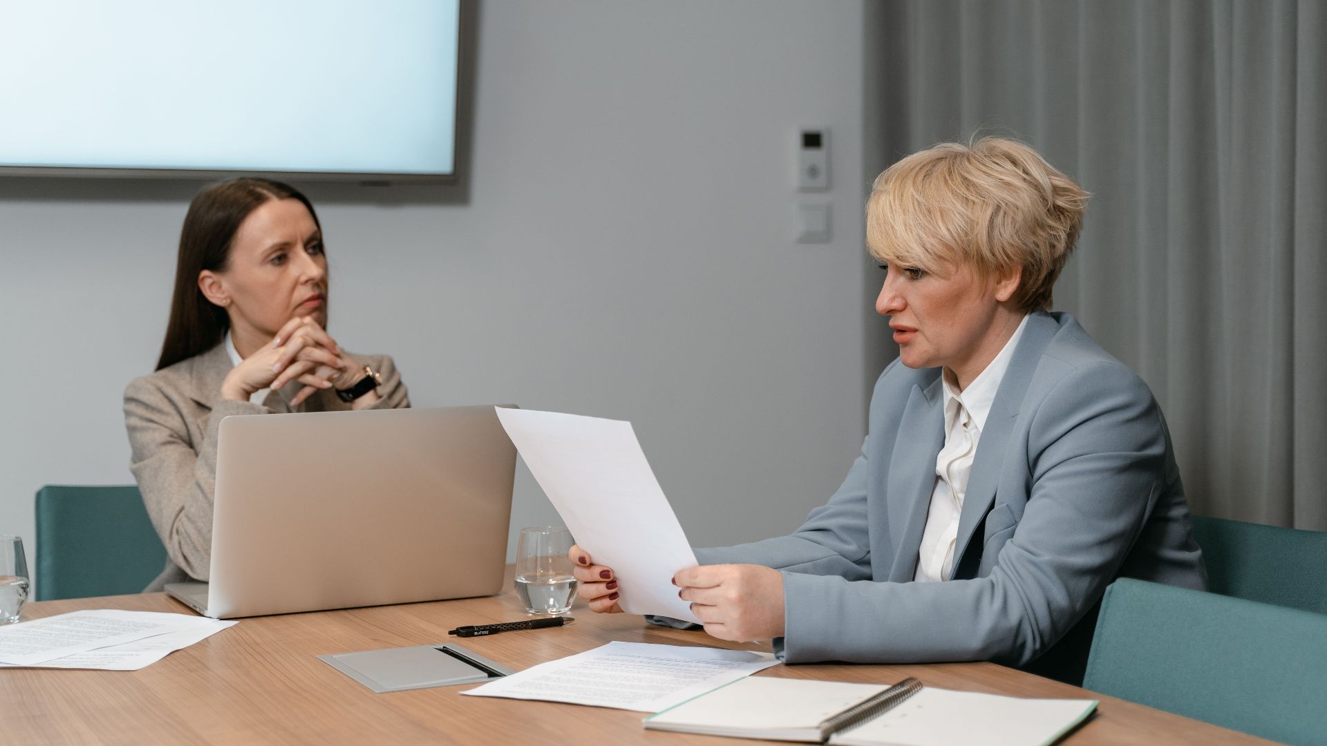 Two businesswomen in a serious meeting, discussing documents with a laptop in an office setting.
