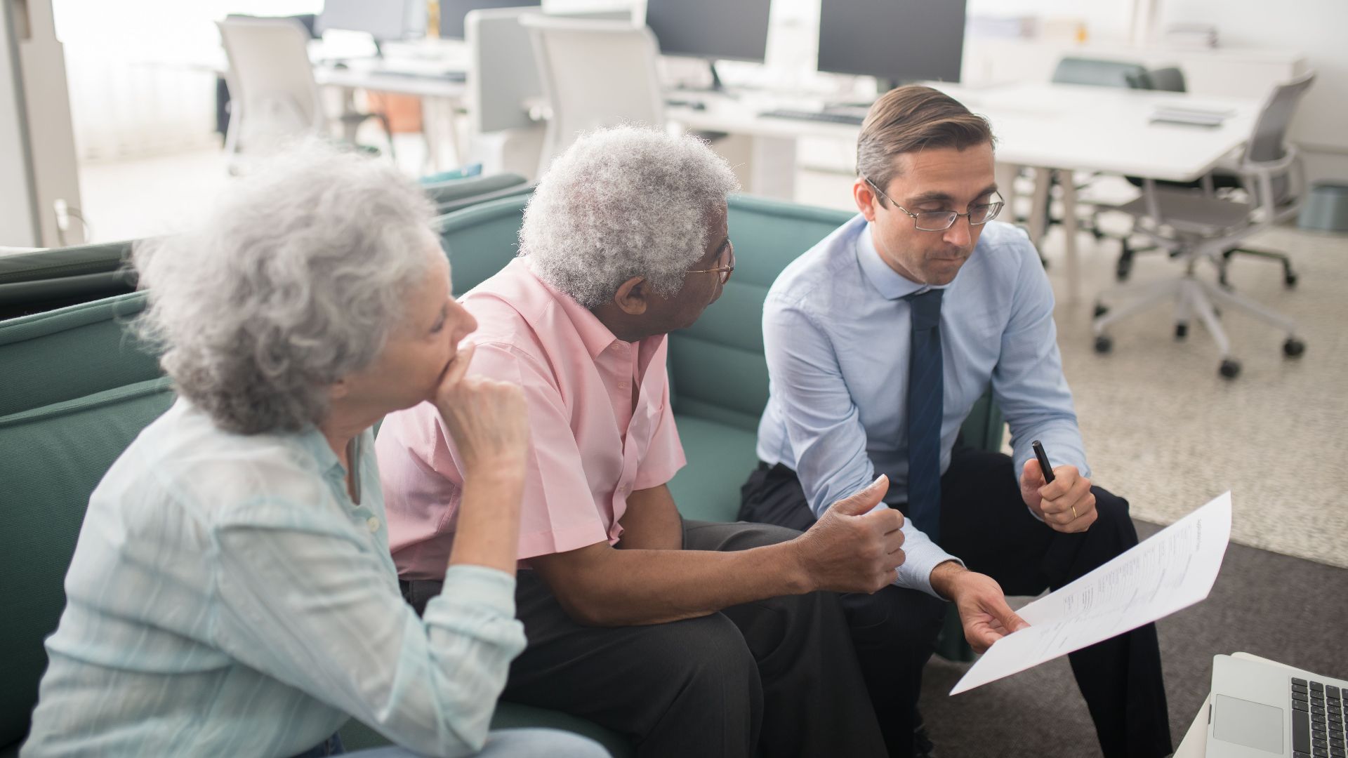 Business professional consults elderly clients in an office setting. Collaborative discussion, paperwork visible.