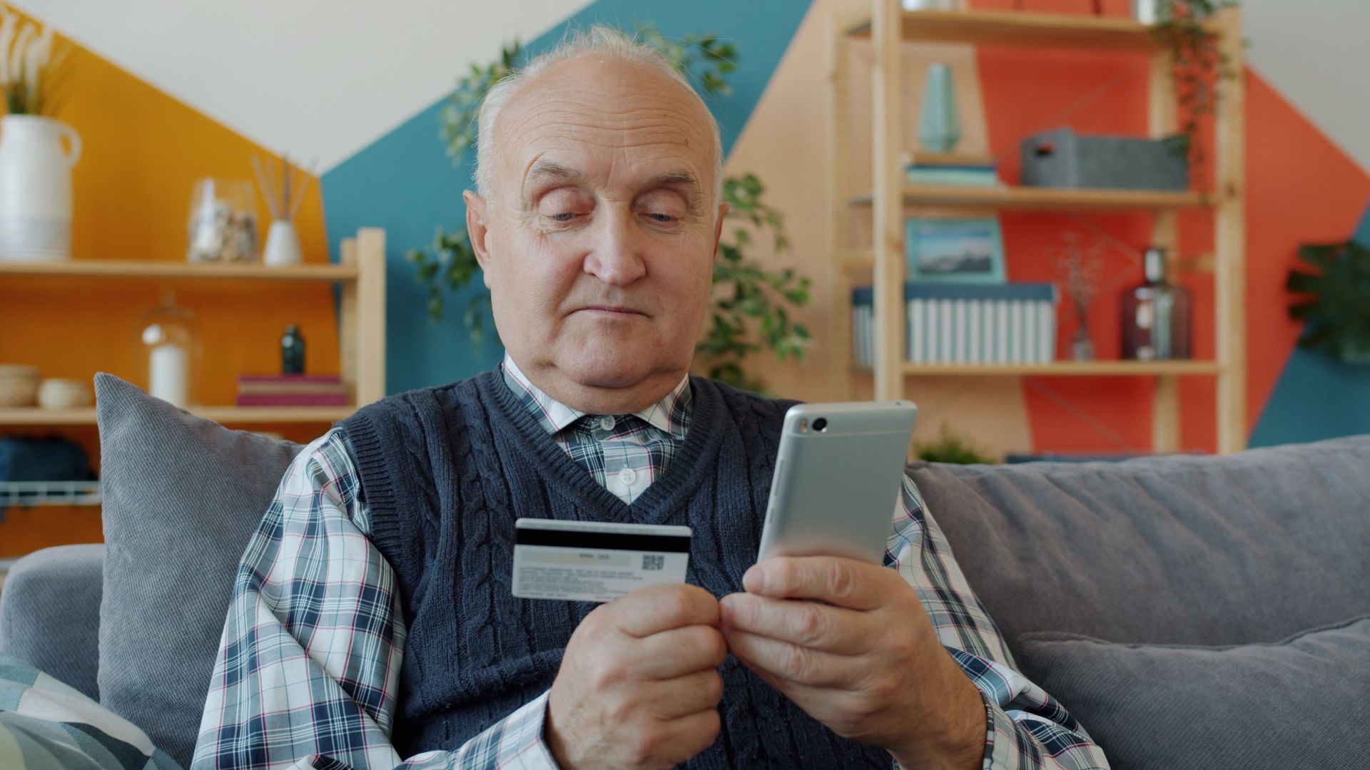 Elderly man using credit card for online shopping with smartphone on couch.