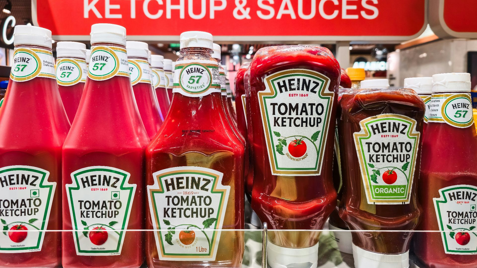 Rows of Heinz tomato ketchup bottles on a supermarket shelf under a red sign