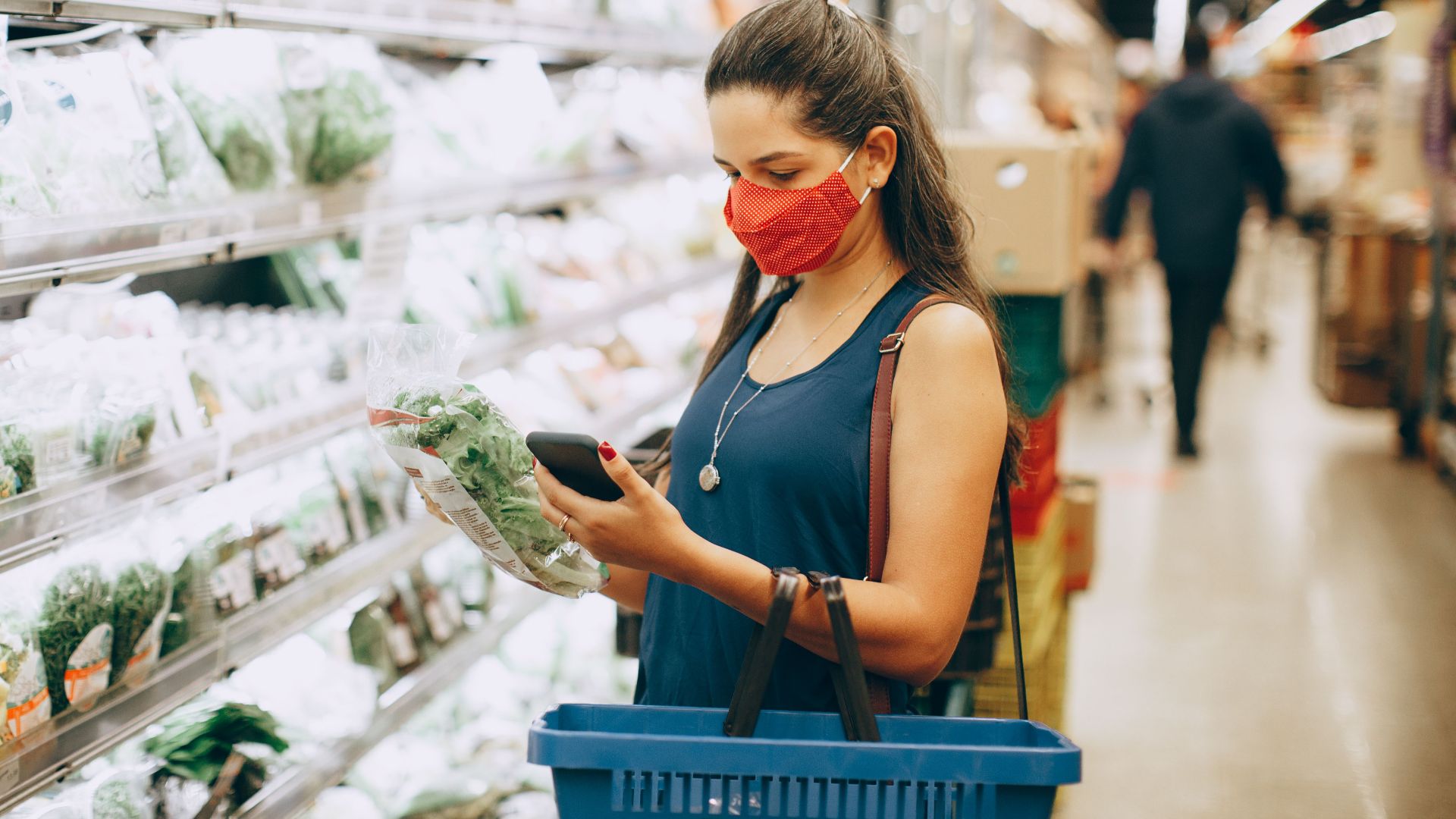 A woman wearing a mask shops for groceries in a supermarket, checking products with her phone.