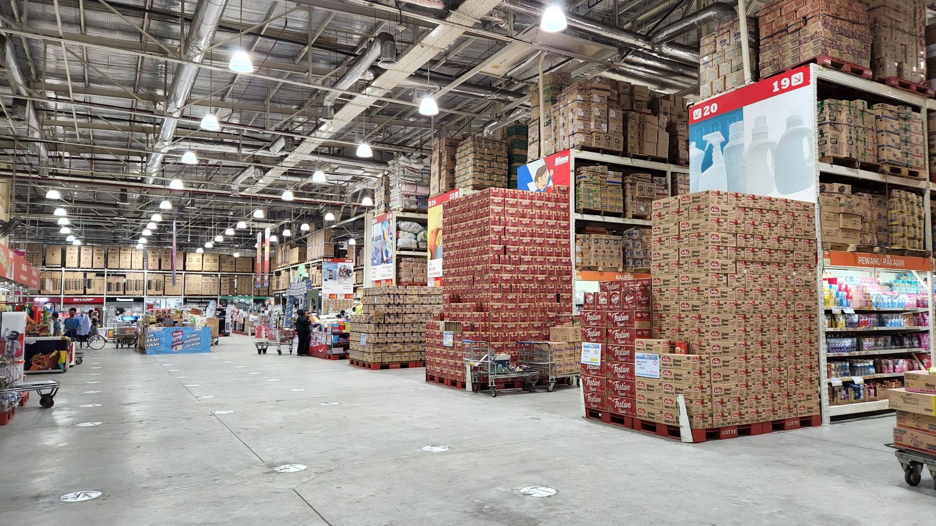 A large wholesale warehouse interior filled with stacked merchandise and shoppers browsing the aisles.