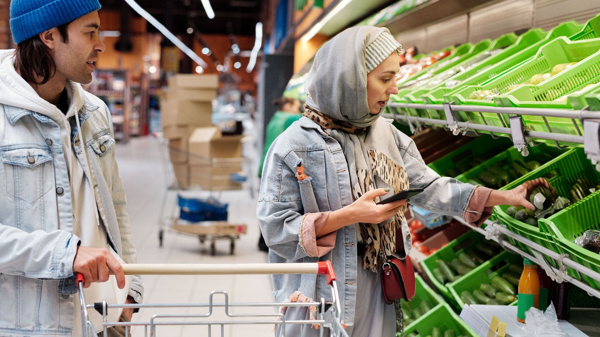 Diverse couple shopping for vegetables in a supermarket, discussing choices with smartphone.
