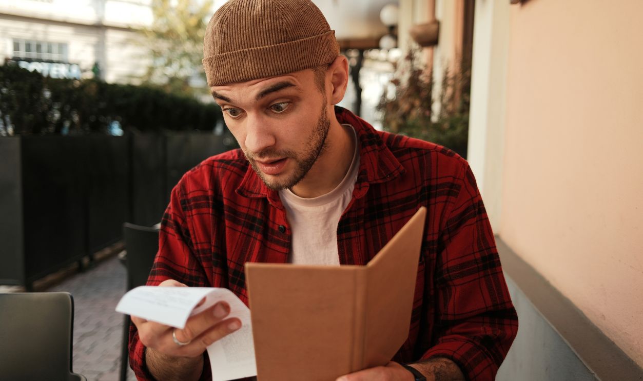 Shocked man holding a restaurant bill and menu while sitting at outdoor cafe