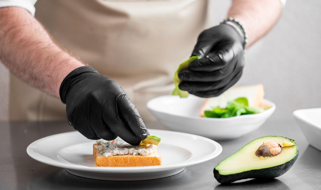 A male chef wearing black nitrile gloves carefully places fresh avocado slices onto a gourmet tuna salad sandwich on a white plate in a professional kitchen setting