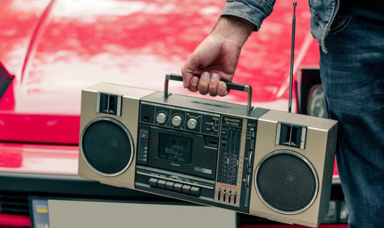 A man holding a vintage cassette player in front of a red classic car, capturing retro vibes