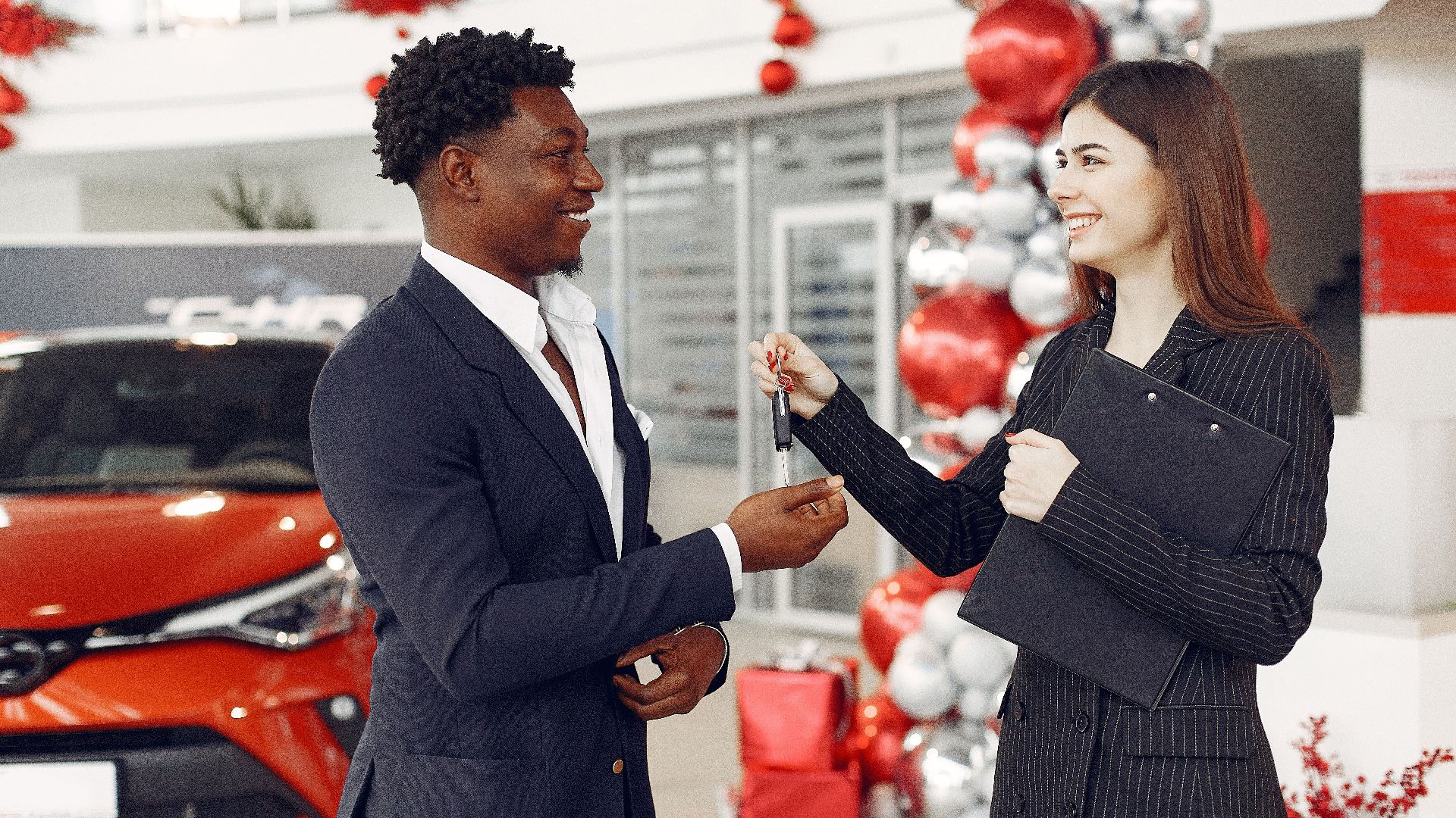 Side view of happy positive female dealer in formal classy wear passing car keys to excited African American customer in trendy suit in car showroom
