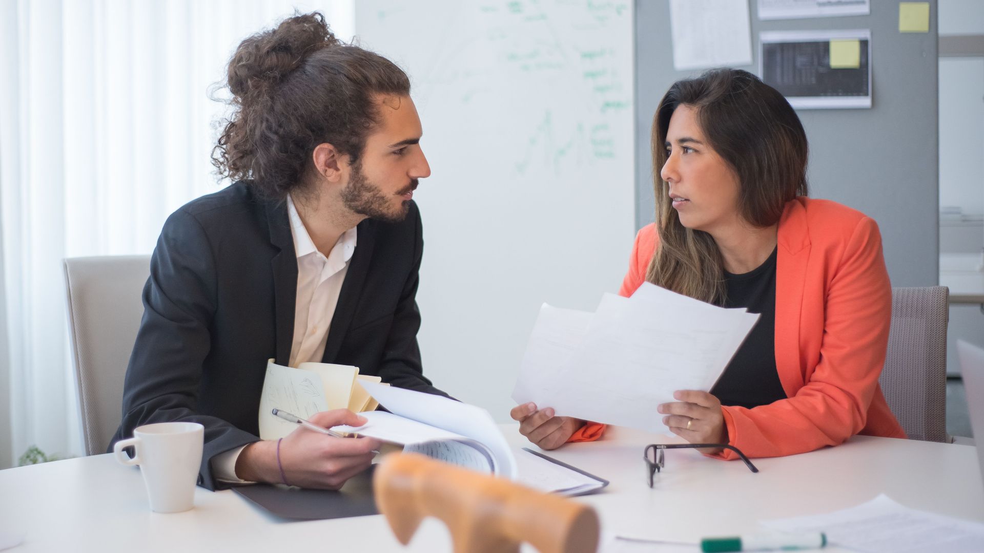 Two colleagues engaged in a discussion at an office table with documents and coffee.