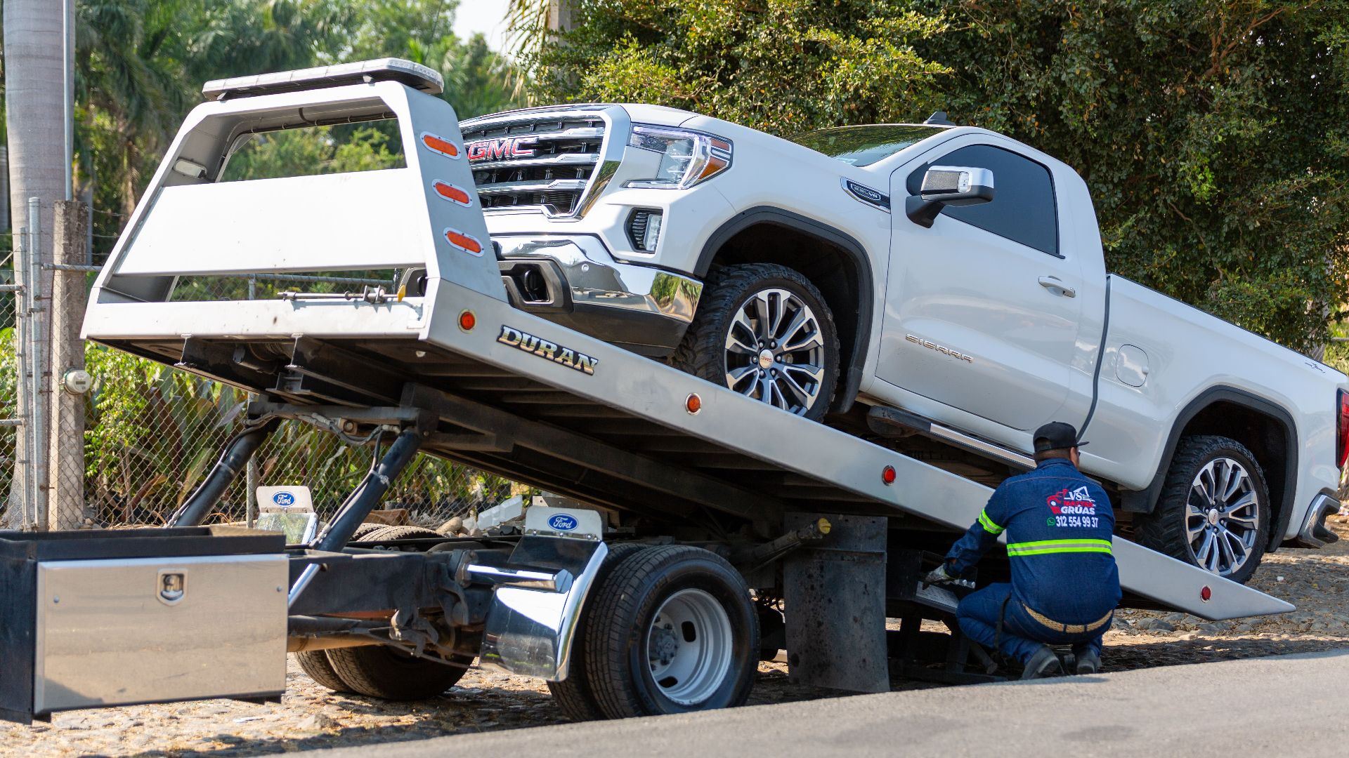 Tow truck operator loading white GMC pickup truck on street in daytime.