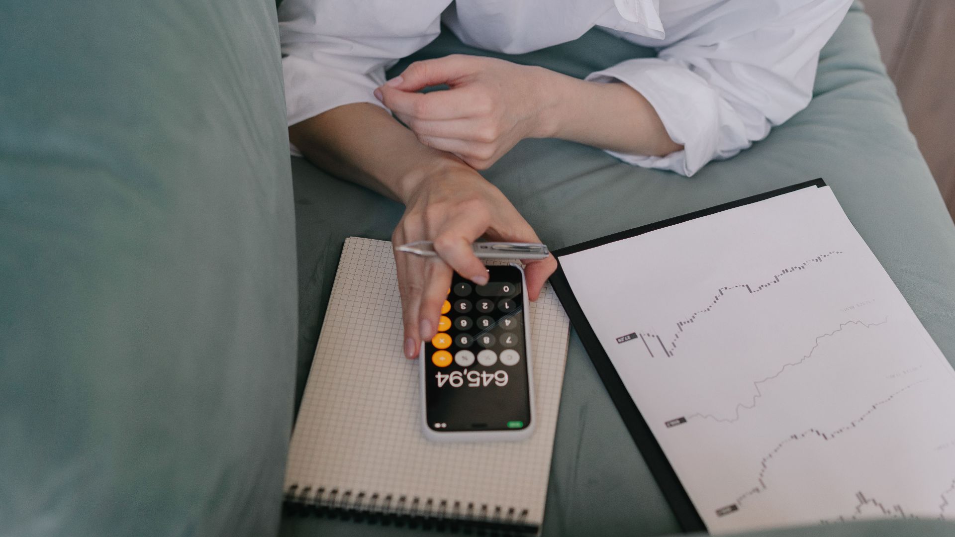 A woman works indoors using a smartphone calculator and analyzing financial graphs, symbolizing modern finance.