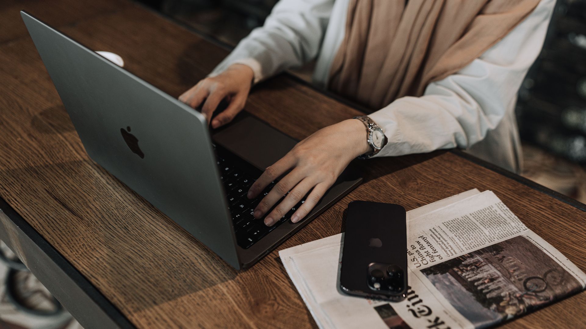Woman typing on a laptop, with a smartphone and newspaper nearby, in a cozy indoor setting.