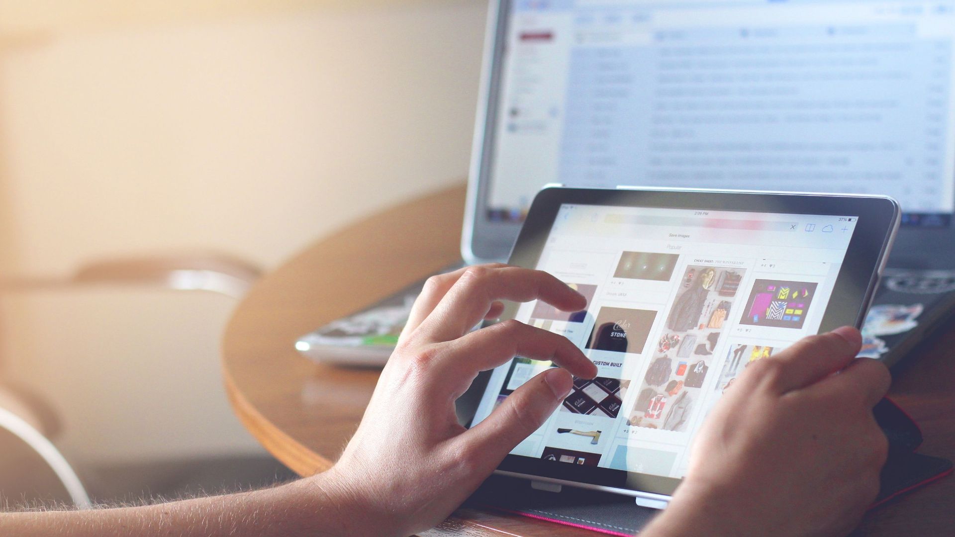 Hands using a tablet beside a laptop in a home office setting, showcasing online browsing.